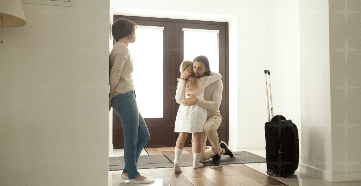 A parent hugging a child at the door of a home. Another person, presumably the mother, looks on forlornly. A packed suitcase is by the door.