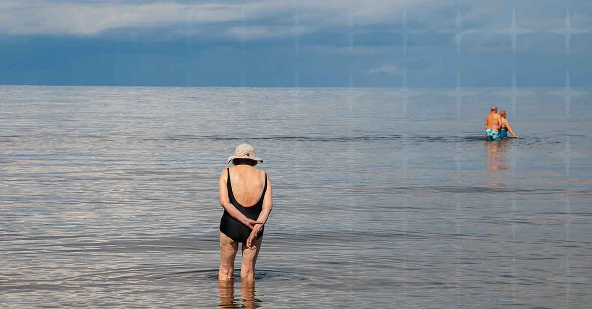 A woman stands at the edge of the sea, watching two of her friends together, who have waded further out into the sea.